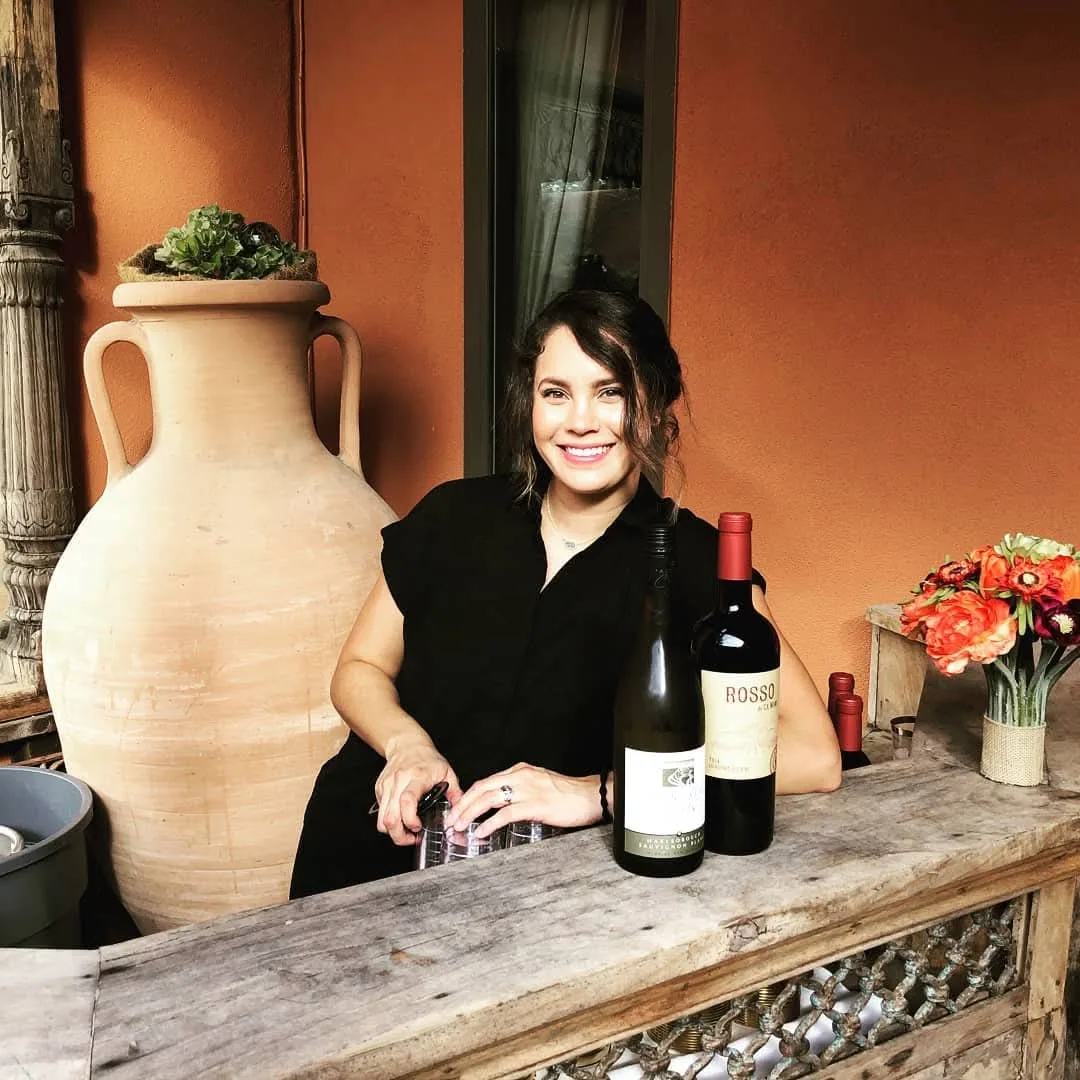 Bar Divas bartender smiling behind a rustic wooden bar with wine bottles and flowers